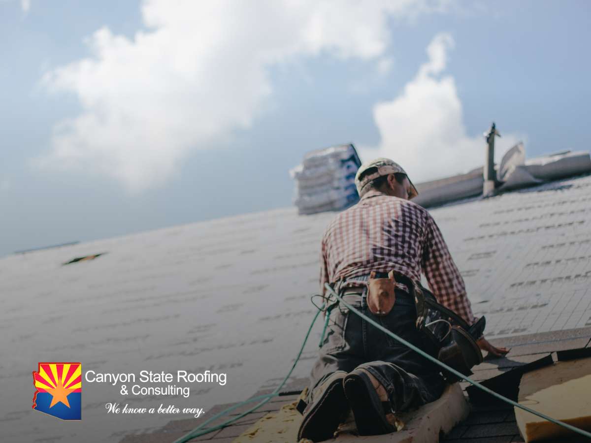 A Phoenix roofer in a checkered shirt installs shingles on a steep roof, secured by a safety rope, with a cloudy sky above.